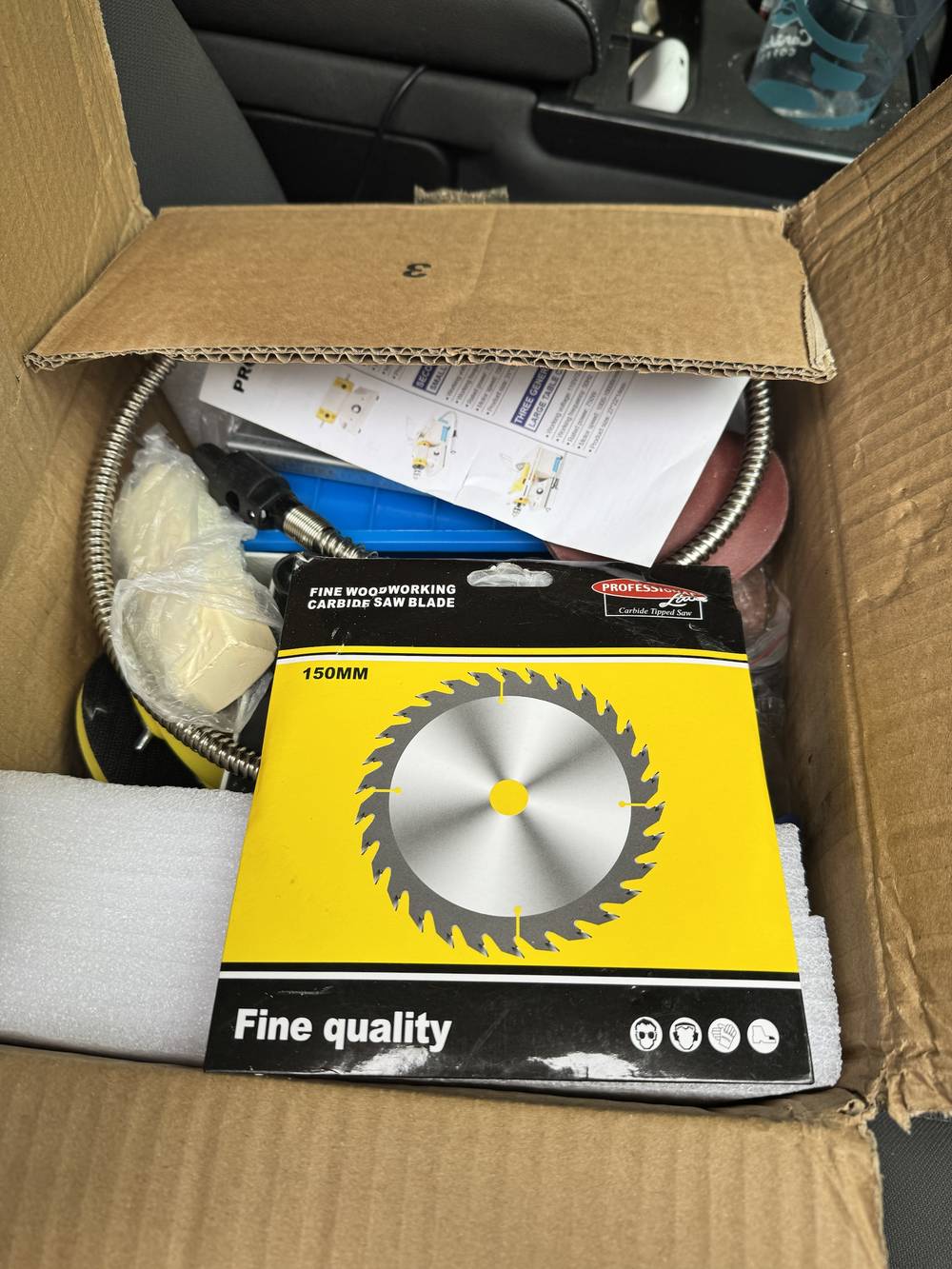 A cardboard box open on a car seat. On top is a blade in a yellow and black packet reading Fine Woodworking Carbide Saw Blade, 150 mm, with a picture of a toothed circular blade. Underneath, a coiled flexible shaft, sanding discs, polishing compound in plastic, and the folded product sheet