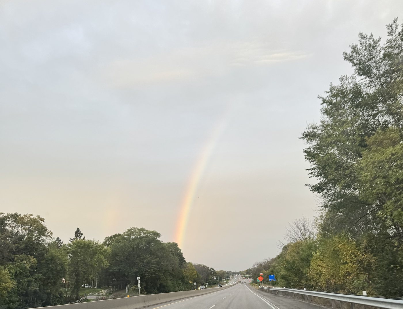A rainbow standing over a divided highway lined with trees under a grey early morning sky, with a faint second bow visible to its left above the trees