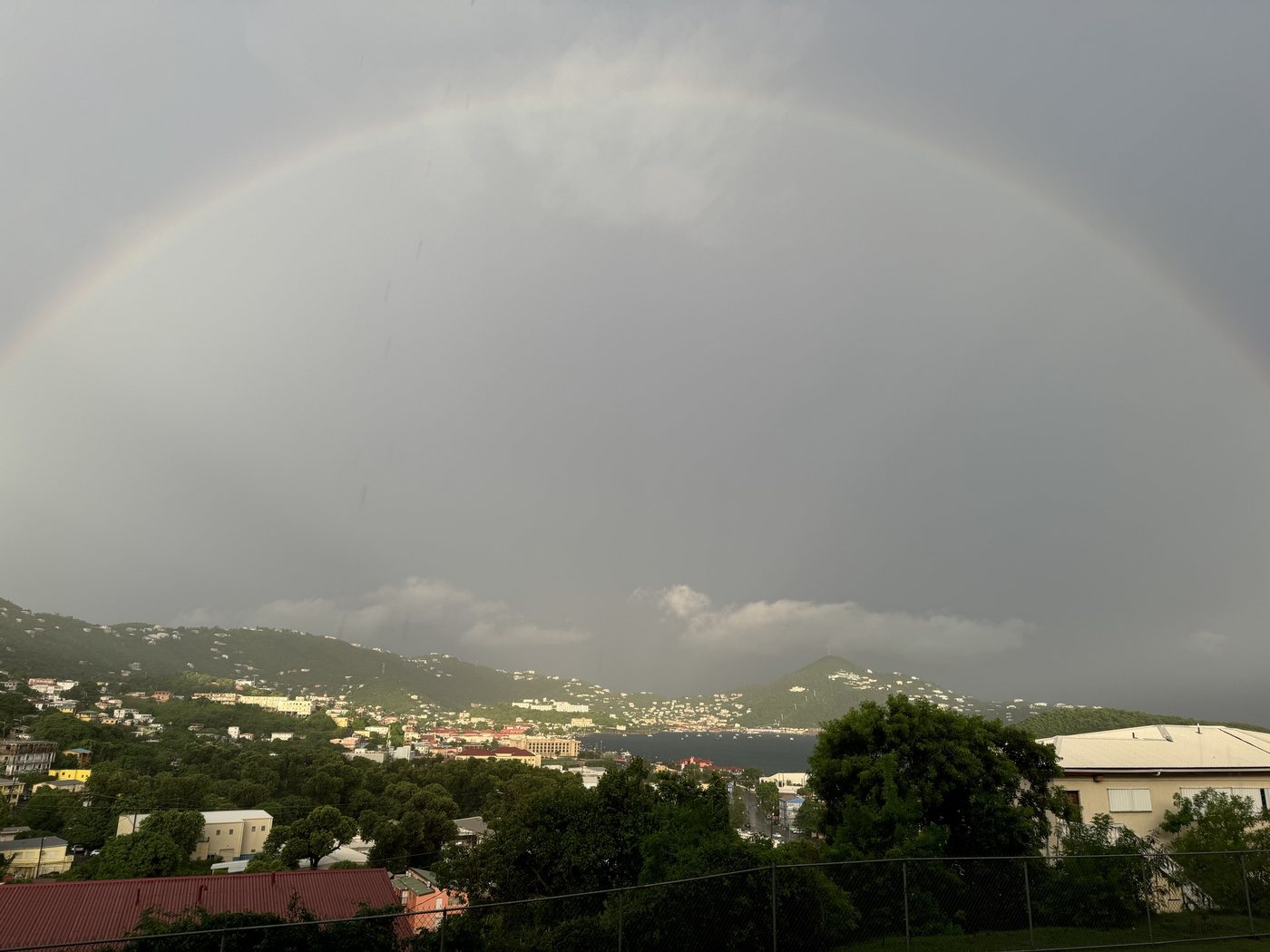 A rainbow arching across the whole sky over a hillside harbour town and a bay, under heavy grey cloud, the arch running off both sides of the frame.