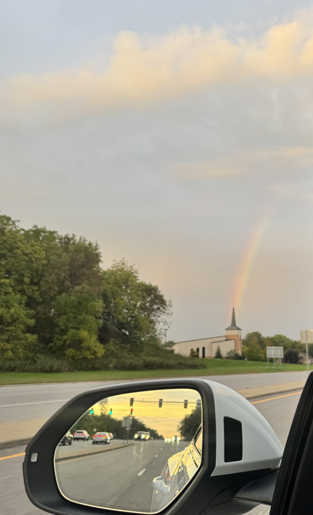 A rainbow coming down behind a church steeple, trees and a road in the foreground, and in the car's side mirror the rising sun glowing gold behind