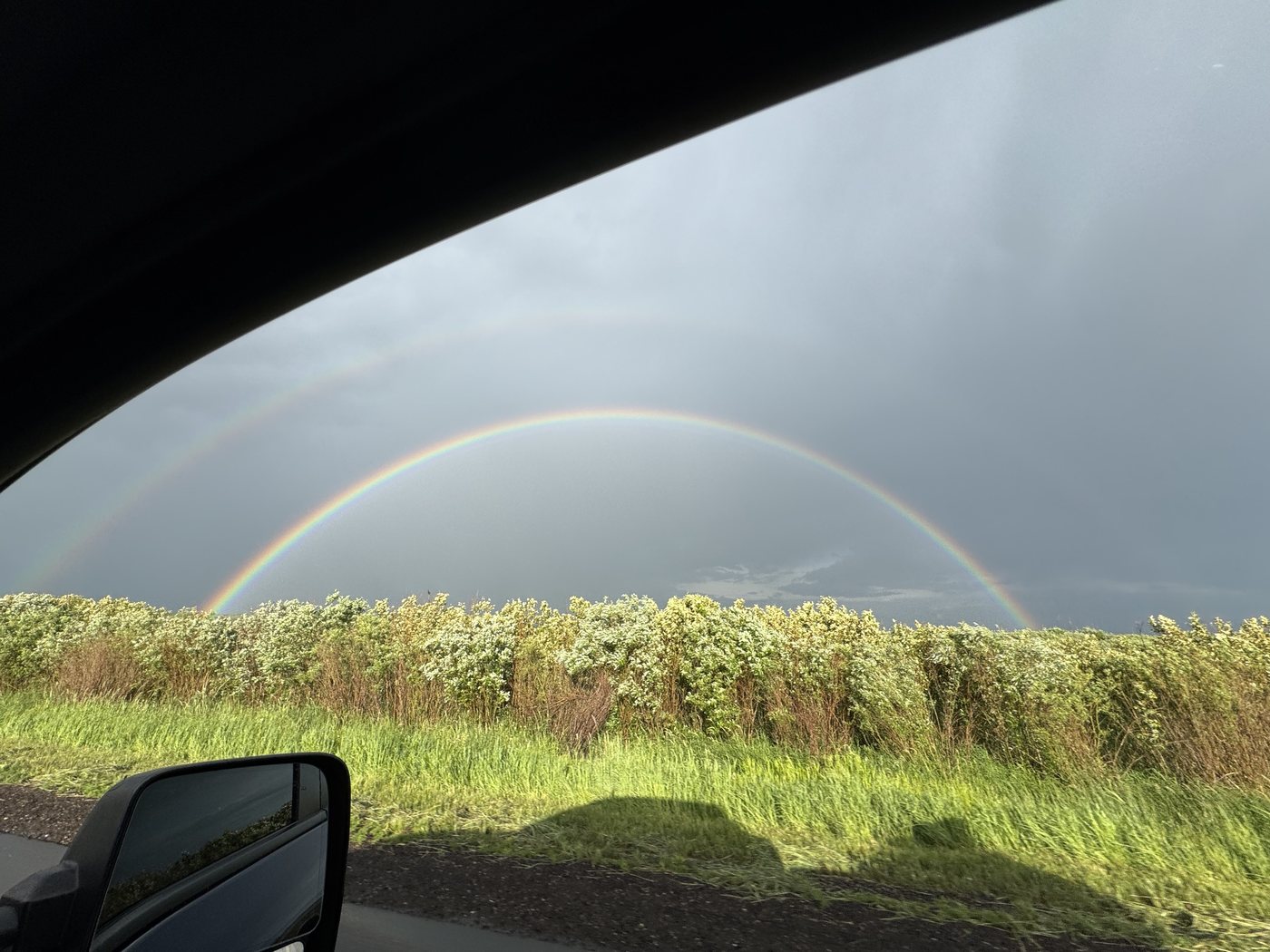 A double rainbow seen through a car window over a line of shrubs, with the car's shadow falling across the grass in the foreground, below the middle of the arch.