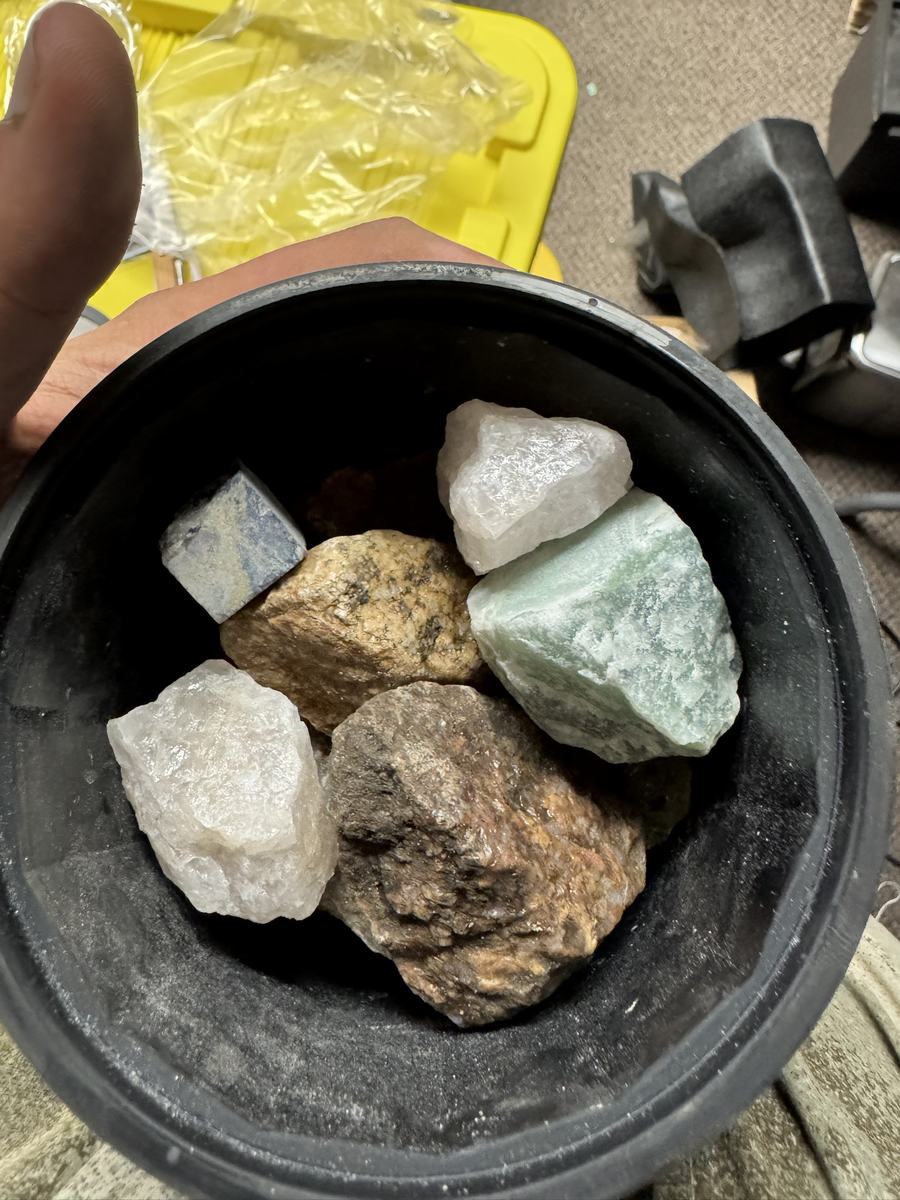 A black rubber tumbler barrel held in one hand, containing five or six rough stones, dry and unpolished. They vary in colour from white and translucent through pale green to tan and grey brown, with visible crystalline texture on the broken surfaces