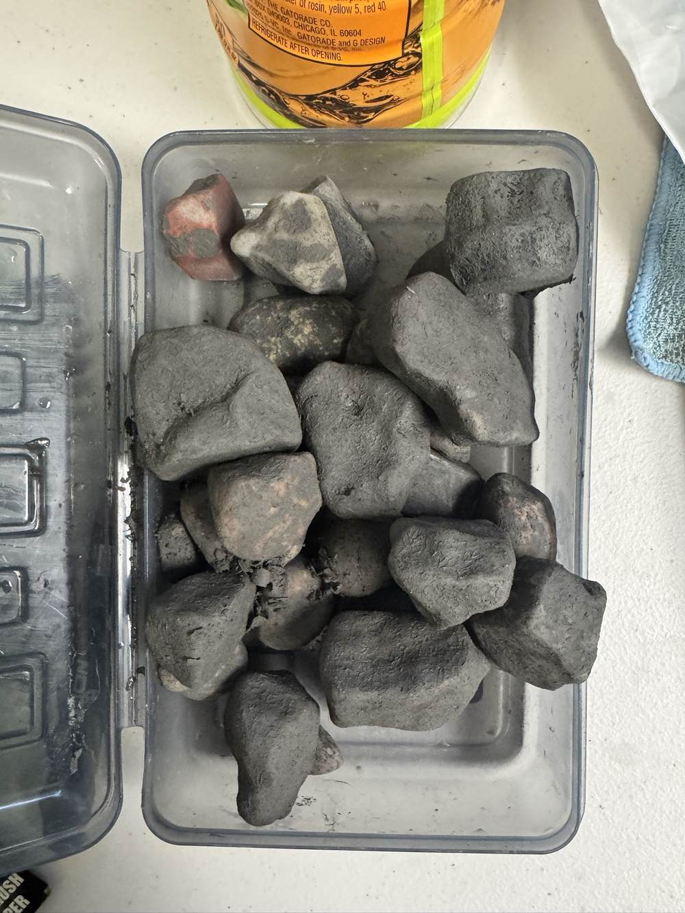A plastic tub holding about sixteen stones, all of them uniformly dull grey and matte, with barely any difference in colour or texture between them. One shows a patch of pink underneath and another a pale streak, but the rest are flatly coated