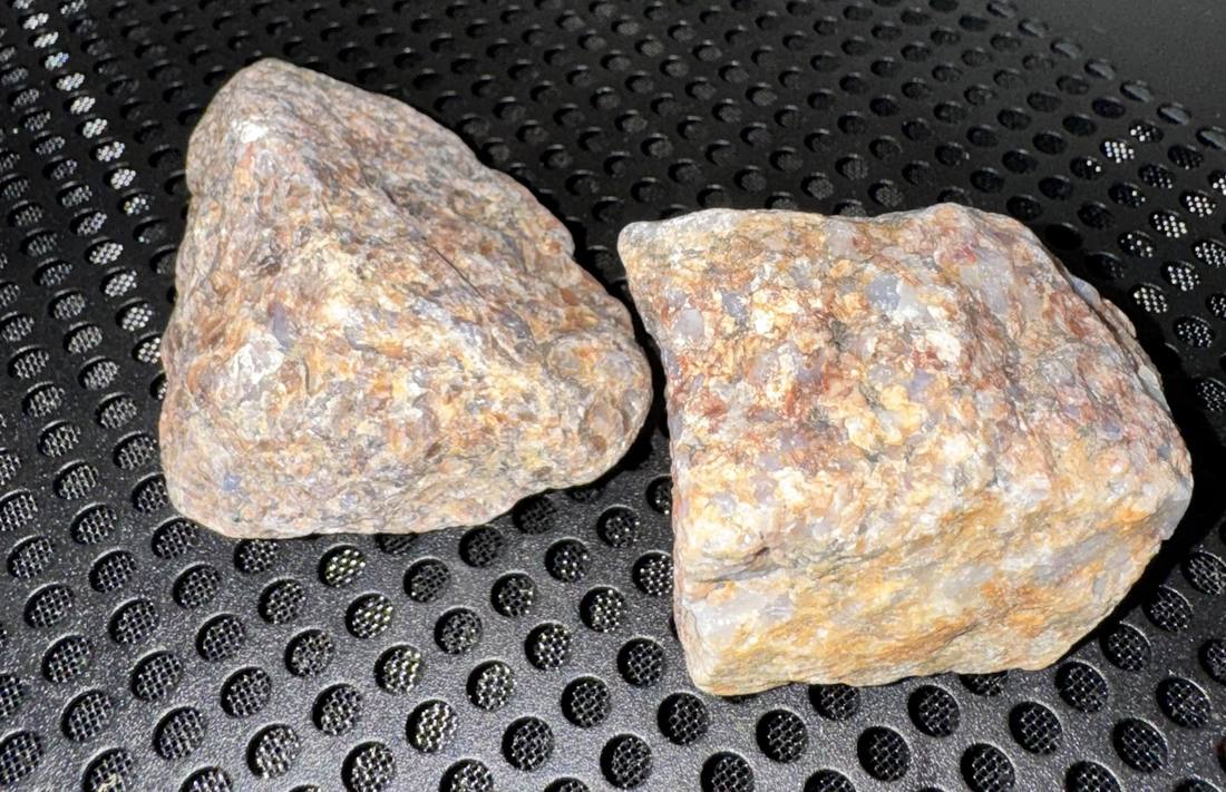 Two rough stones photographed dry on a black perforated surface. Both are pale, mottled in orange, cream and grey, with translucent patches and a waxy looking sheen on the broken faces. The nearer one is roughly wedge shaped, the other more angular