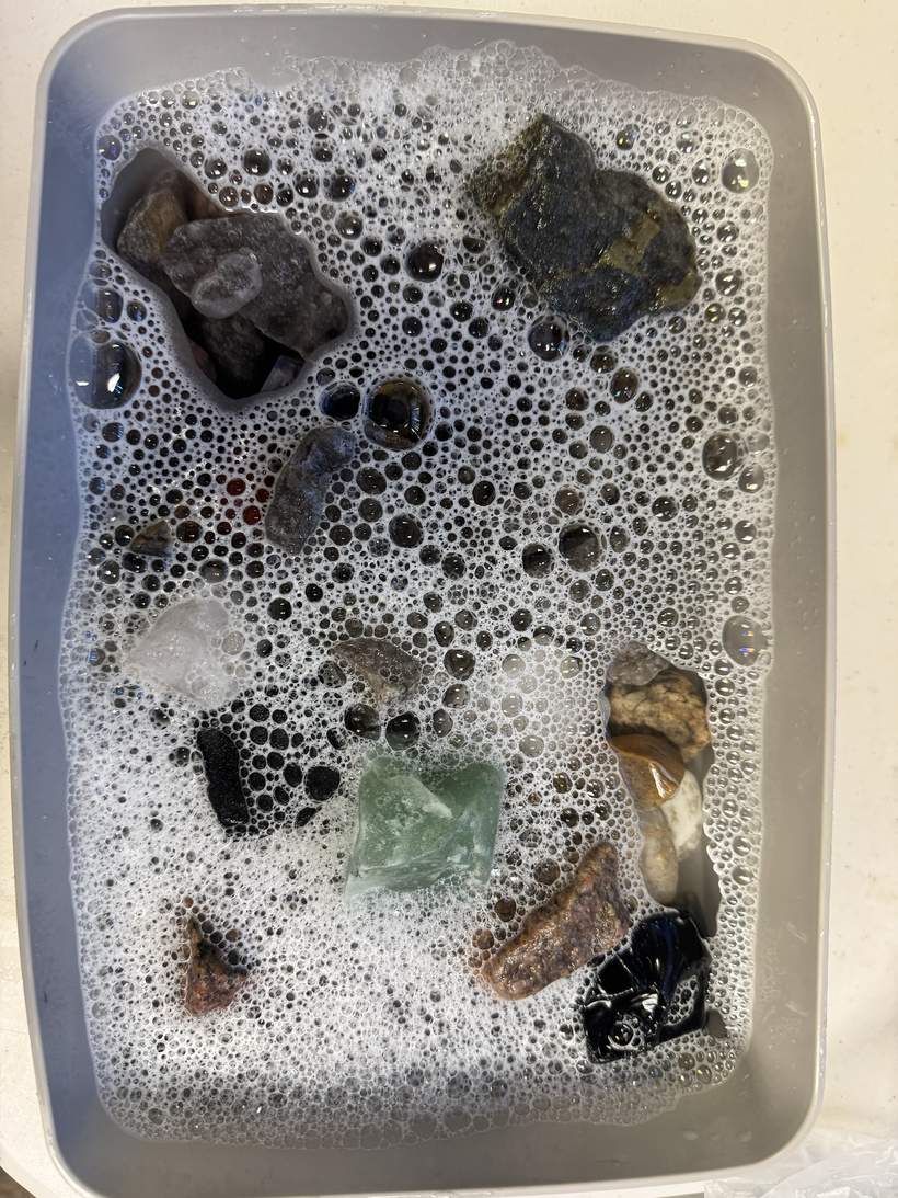 A grey plastic tub of stones soaking in soapy water, seen from above, the surface covered in foam and bubbles of varying size. A dozen or so rough stones of different colours are visible through and around the foam, some dark, some pale and translucent, one green and angular near the centre